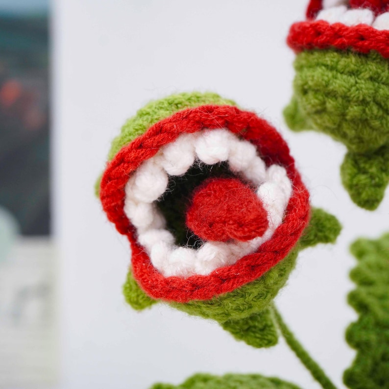 May include: Crocheted green plant with a red-lined mouth filled with white teeth and a red tongue. The plant has a whimsical, cartoonish appearance, with a vibrant color palette and textured yarn. The plant's design is reminiscent of a carnivorous plant.