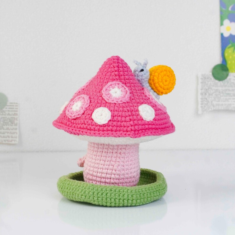 May include: A handmade crochet mushroom with a pink cap, white polka dots, and a light pink stem. A small snail with a yellow shell sits on top. The mushroom is set on a green base, creating a whimsical, decorative piece.