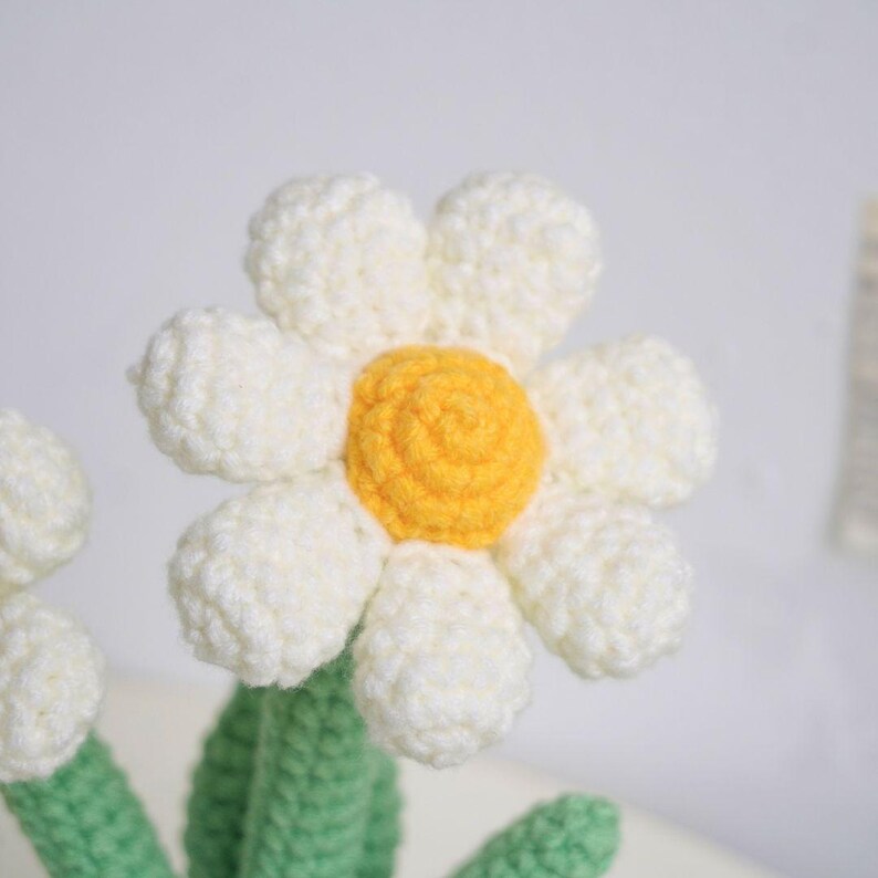 Puede incluir: Una flor de margarita de crochet con un centro blanco y un centro amarillo. La flor es parte de un ramo de flores de crochet.