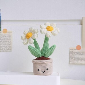 Puede incluir: Una planta de margarita de crochet con una maceta sonriente. La planta tiene dos margaritas blancas con centros amarillos y hojas verdes. La maceta es de color marr&oacute;n claro.