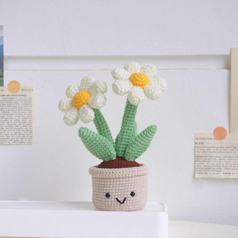Puede incluir: Una planta de margarita de crochet con una maceta sonriente. La planta tiene dos margaritas blancas con centros amarillos y hojas verdes. La maceta es de color marr&oacute;n claro.