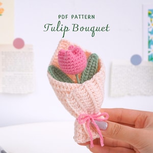 以下が含まれることがあります： ピンクの毛糸で編まれたチューリップのブーケが、ピンクの毛糸で編まれた紙に包まれています。ブーケはピンクのリボンで結ばれています。画像の上部には「PDF PATTERN Tulip Bouquet」というテキストがあります。