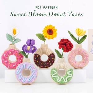 Può includere: Sei vasi all'uncinetto a forma di ciambella, ognuno con un diverso fiore all'uncinetto. I vasi sono in vari colori, tra cui rosa, marrone e verde, con decorazioni a spruzzo. Il testo "Sweet Bloom Donut Vases" è in alto.