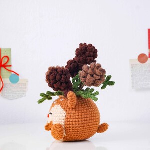 May include: A handmade crochet deer-shaped vase in shades of brown, tan, and white, holding several brown pinecones and green leaves. The deer has small ears, a white face, and a red nose. The vase is a unique decorative item.