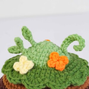 May include: Crocheted hat with a whimsical design. The hat features a green, textured crown with leaf-like details and decorative orange and yellow flower accents. The base of the hat is a mix of brown and green.