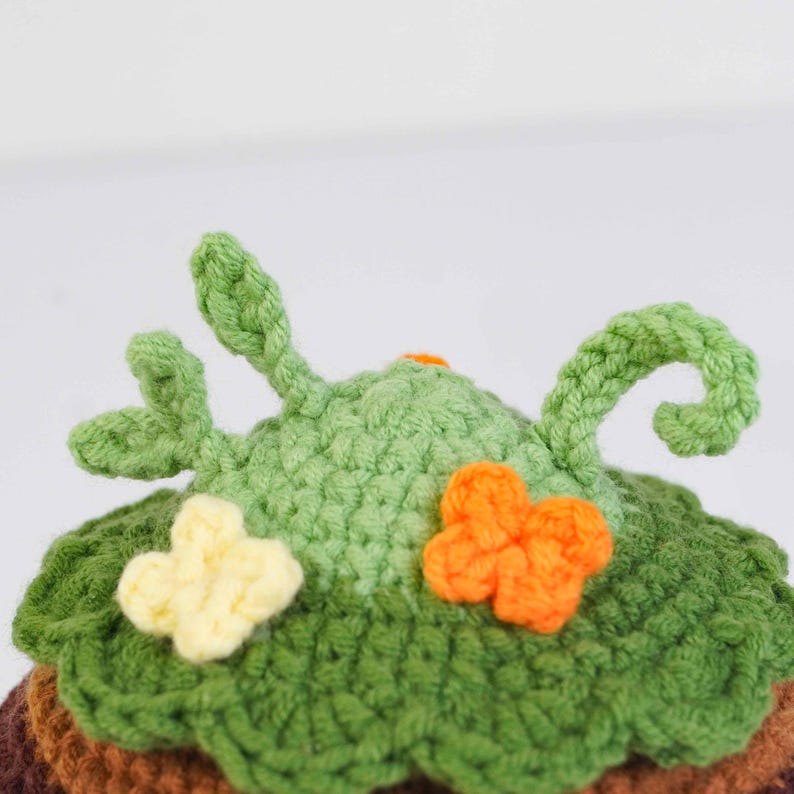 May include: Crocheted hat with a whimsical design. The hat features a green, textured crown with leaf-like details and decorative orange and yellow flower accents. The base of the hat is a mix of brown and green.