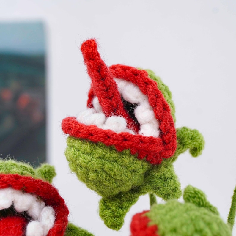 May include: Crocheted green plant-like figures with red-lined mouths and white teeth. The figures have a red tongue and are set against a soft background. The crochet work is detailed, with visible stitches.