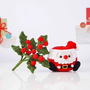 May include: A crocheted Christmas decoration featuring a Santa Claus figure and a sprig of holly. The Santa Claus is in a seated position, with a red hat and suit, white beard, and black boots. The holly has green leaves and red berries.