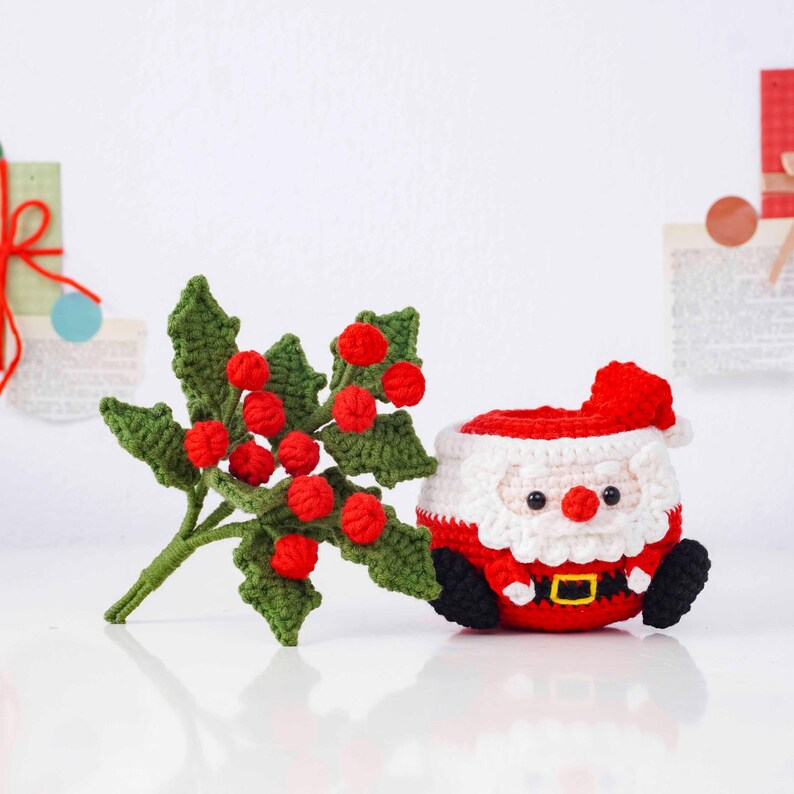 May include: A crocheted Christmas decoration featuring a Santa Claus figure and a sprig of holly. The Santa Claus is in a seated position, with a red hat and suit, white beard, and black boots. The holly has green leaves and red berries.