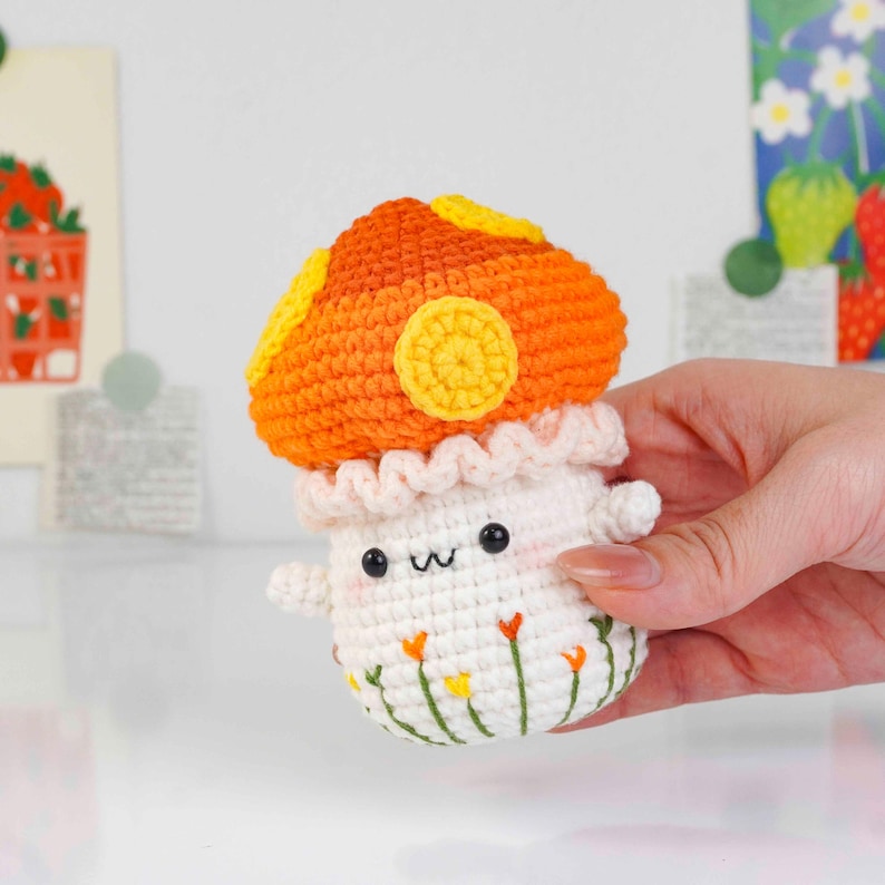 May include: A handmade crochet mushroom toy with an orange cap and yellow accents. The mushroom has a white stem with a smiling face and small arms. The base is decorated with embroidered flowers. A whimsical, soft toy.