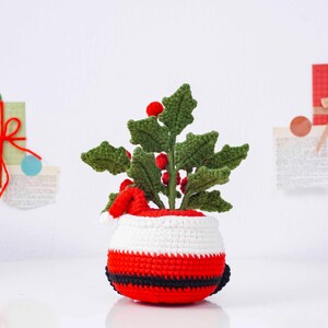 May include: A crocheted Christmas decoration featuring a holly plant with green leaves and red berries. The plant is in a pot designed to look like Santa Claus, with red, white, and black stripes and a small Santa hat.