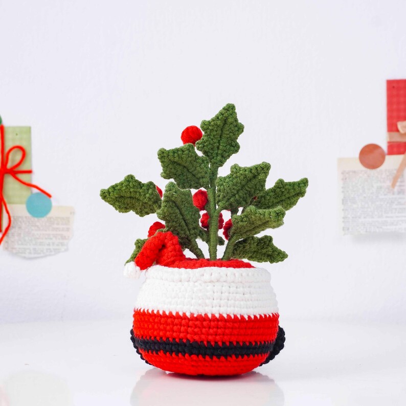 May include: A crocheted Christmas decoration featuring a holly plant with green leaves and red berries. The plant is in a pot designed to look like Santa Claus, with red, white, and black stripes and a small Santa hat.