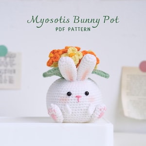 Può includere: Un vaso a forma di coniglio bianco all'uncinetto con fiori arancioni e gialli in cima. Il vaso è a forma di coniglio con un corpo rotondo, orecchie lunghe e un piccolo naso. I fiori sono disposti in un cerchio attorno alla testa del coniglio. Il testo "Myosotis Bunny Pot PDF Pattern" è scritto sopra il coniglio.