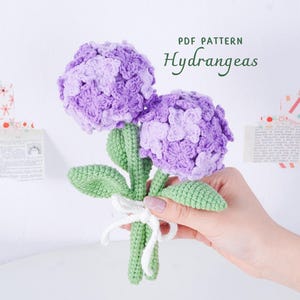 Puede incluir: Un ramo de hortensias hecho a mano con dos grandes flores moradas. El tallo y las hojas verdes están detallados, y el ramo está atado con una cinta blanca. El texto "PDF PATTERN Hydrangeas" es visible.