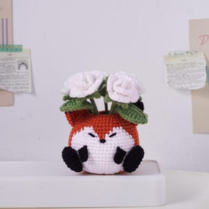 May include: A handcrafted crochet fox planter in orange, white, and black, holding white crochet flowers with green leaves. The fox planter has a smiling face and black paws. A charming decorative piece for home decor.