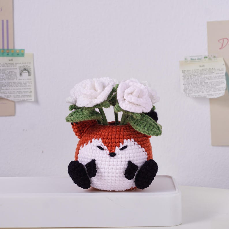 May include: A handcrafted crochet fox planter in orange, white, and black, holding white crochet flowers with green leaves. The fox planter has a smiling face and black paws. A charming decorative piece for home decor.