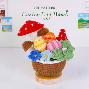 Puede incluir: Un cuenco de huevos de Pascua tejido a ganchillo con un diseño de seta y huevos. El cuenco marrón tiene un borde blanco, con huevos coloridos, flores azules y hojas verdes. La seta tiene un gorro rojo con lunares blancos. Texto: "PDF PATTERN Easter Egg Bowl."