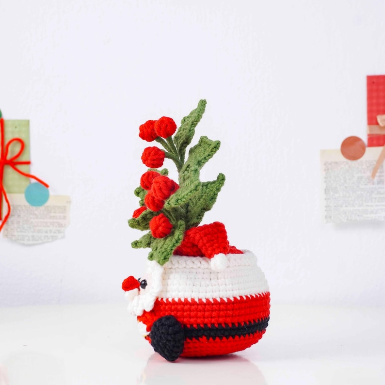 May include: A crocheted Santa Claus planter with a red and white body, black belt, and a small red hat. The planter holds a sprig of green leaves and red berries, creating a festive holiday decoration.
