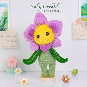 May include: A crocheted flower doll with a yellow centre, purple petals, and green stem and leaves. The doll stands on a wooden base. The text "Baby Orchid PDF Pattern" is above the doll.