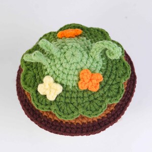 May include: A handmade crochet cake with green frosting, decorated with orange and yellow flower accents. The cake has a brown base and a light brown middle layer. The top layer is green, with a light green frog design.