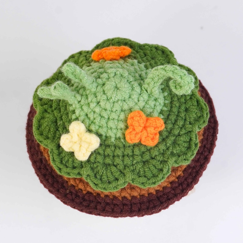 May include: A handmade crochet cake with green frosting, decorated with orange and yellow flower accents. The cake has a brown base and a light brown middle layer. The top layer is green, with a light green frog design.