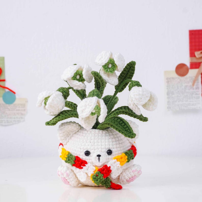 May include: A handmade crochet flower pot shaped like a white bear, adorned with a colorful scarf. The pot holds a bouquet of white and green crochet flowers. The bear has a black nose and small pink paws.