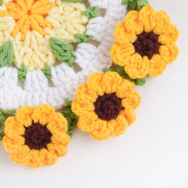 May include: Close-up of a handmade crochet doily featuring a sunflower design. The doily has a white, yellow, and green color scheme. The sunflowers are yellow with brown centers. The leaves are green.