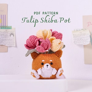 以下が含まれることがあります： ピンクと黄色のチューリップが入った、かぎ針編みの柴犬ポット。 ポットはオレンジ色で白いアクセントがあり、漫画のようなデザインです。 テキスト「PDF PATTERN Tulip Shiba Pot」がポットの上に表示されています。