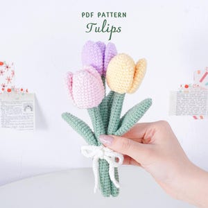 May include: A handmade bouquet of crocheted tulips in pink, lilac, and yellow, with green stems and leaves. The flowers are tied with a white ribbon. The text "PDF PATTERN Tulips" is at the top.