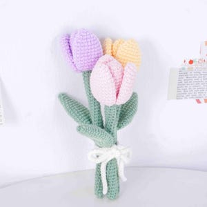 May include: A bunch of three crocheted tulips in shades of purple, pink, and yellow. The flowers have green stems and leaves, tied with a white crocheted bow. These handmade flowers are a charming decorative item.