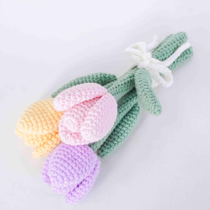 May include: A handmade crochet bouquet of tulips in pastel shades. The bouquet features three tulips in yellow, pink, and purple, with green leaves and stems. A white crocheted bow secures the stems. The flowers are arranged on a white background.