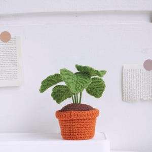 May include: A crocheted plant with green leaves in a brown and orange pot. The plant is sitting on a white surface.