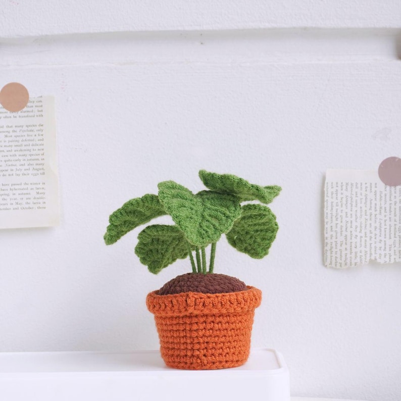 May include: A crocheted plant with green leaves in a brown and orange pot. The plant is sitting on a white surface.