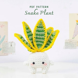 May include: A crocheted snake plant with yellow and green leaves and a white pot. The plant has a cute face with black eyes and pink cheeks. The text "PDF PATTERN Snake Plant" is at the top.