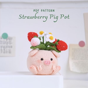 Op de afbeelding: Een gehaakte varkens pot met een roze lichaam, zwarte ogen en een roze snuit. De pot is gevuld met een groene plant met rode en witte gehaakte aardbeien en witte gehaakte bloemen.