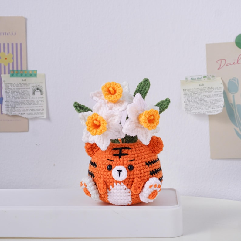 May include: A handmade crochet tiger-shaped flower pot, filled with white and yellow daffodils. The orange and black striped pot has a cute face and paws. A charming, whimsical home decor item.