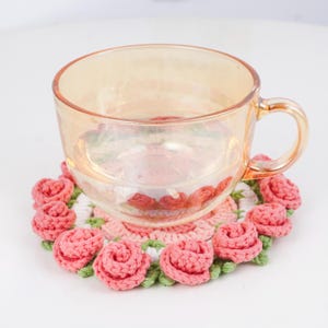 Puede incluir: Una taza de t&eacute; de vidrio transparente descansa sobre un posavasos de ganchillo hecho a mano. El posavasos presenta un dise&ntilde;o de rosas rosas con hojas verdes y un fondo blanco. La taza tiene un tono dorado.