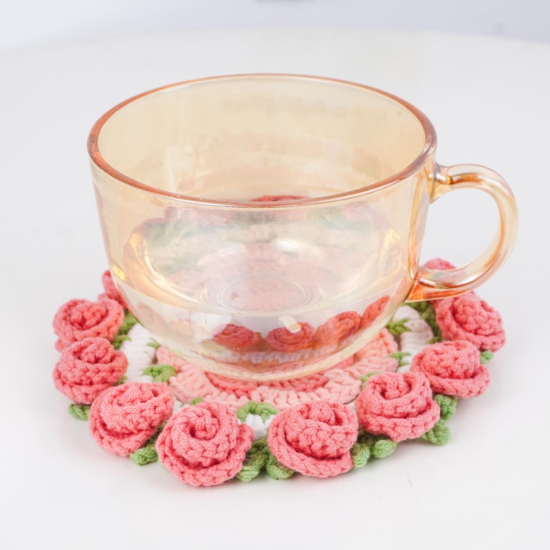 Puede incluir: Una taza de t&eacute; de vidrio transparente descansa sobre un posavasos de ganchillo hecho a mano. El posavasos presenta un dise&ntilde;o de rosas rosas con hojas verdes y un fondo blanco. La taza tiene un tono dorado.