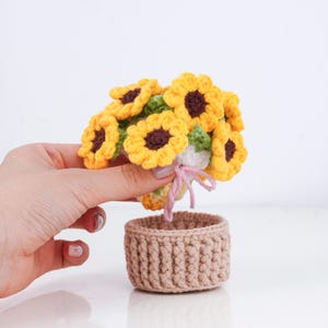 May include: A handmade crochet bouquet of sunflowers with yellow petals and brown centers, held above a small, tan crochet basket. The flowers are detailed with green leaves and tied with a pink ribbon.