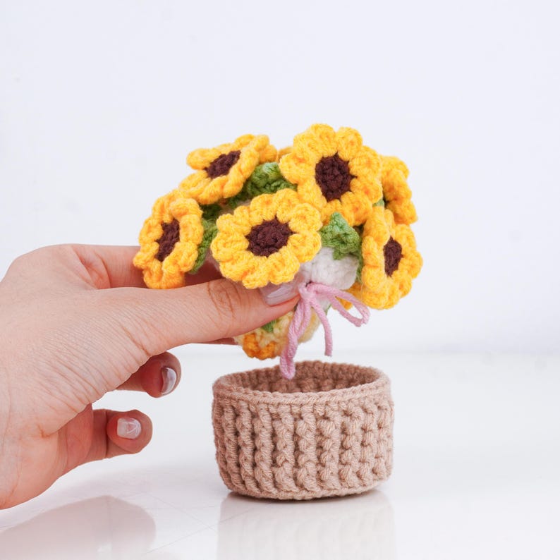 May include: A handmade crochet bouquet of sunflowers with yellow petals and brown centers, held above a small, tan crochet basket. The flowers are detailed with green leaves and tied with a pink ribbon.