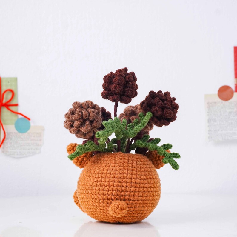 May include: A handmade crochet flower arrangement in a round orange pot. The arrangement features brown and tan flowers with green leaves. The pot has small ear-like details on the sides and a small tail-like detail on the back.