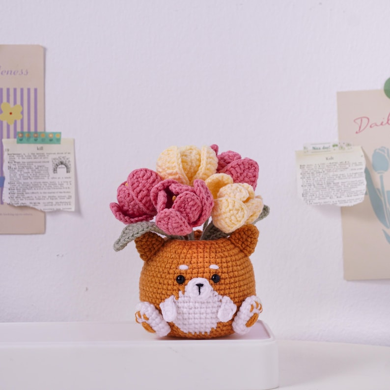 May include: A handmade crochet flower pot shaped like a Shiba Inu dog. The pot is orange and white, with a black nose and eyes. It holds a bouquet of pink and yellow crochet tulips. The pot is approximately 4 inches tall.