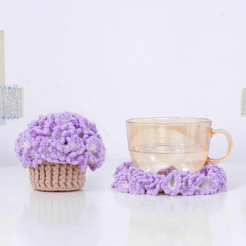 以下が含まれることがあります： ラベンダーの花の形をしたカップカバーと、お揃いのコースターが特徴の手作りのかぎ針編みセット。カップカバーは、ベージュのベースとふわふわのラベンダーのトップが付いたカップケーキのようにデザインされています。コースターはラベンダーの花のリングです。