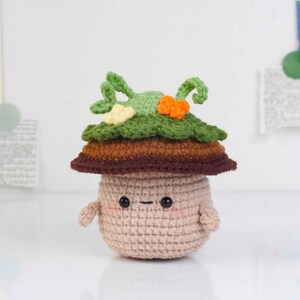 May include: A crocheted mushroom figure with a tan body, brown and green layered cap, and small arms. The mushroom has a smiling face with black eyes and a dash for a mouth. The cap features green leaves and orange and yellow accents.