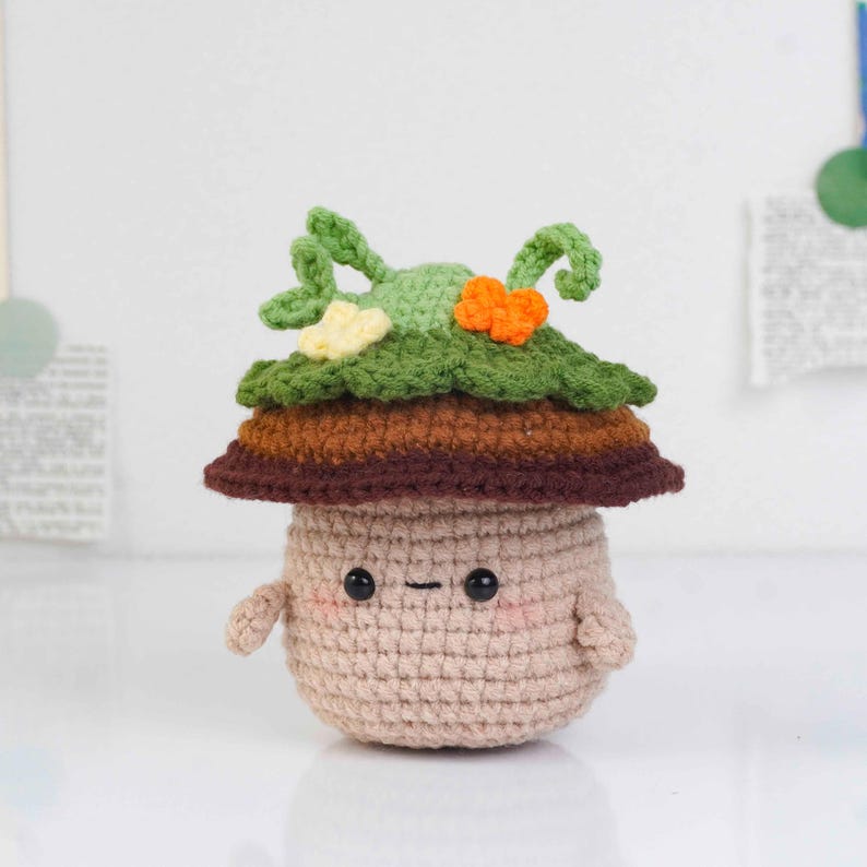 May include: A crocheted mushroom figure with a tan body, brown and green layered cap, and small arms. The mushroom has a smiling face with black eyes and a dash for a mouth. The cap features green leaves and orange and yellow accents.