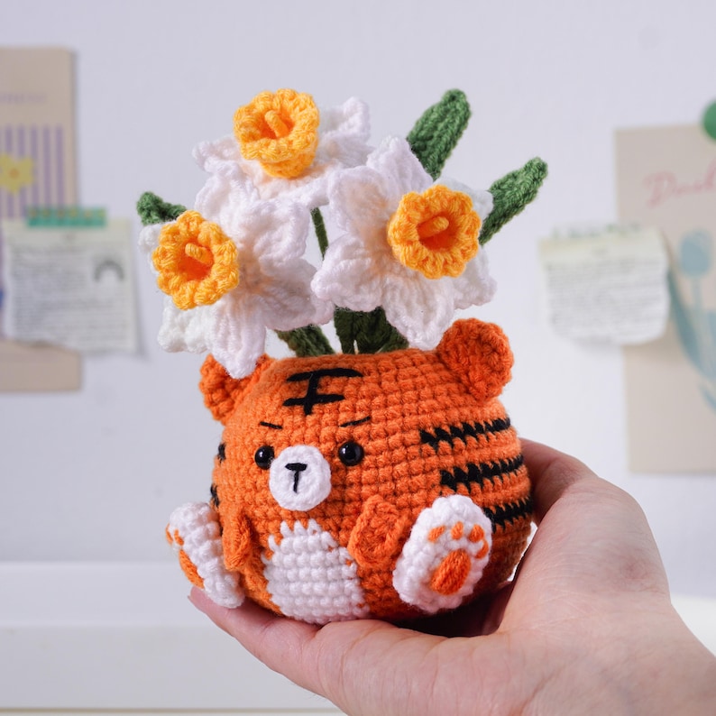 May include: A handmade crochet tiger-shaped flower pot in orange, black, and white, holding white and yellow crochet daffodils. The tiger has black stripes, a black nose, and a white belly and paws. The flowers have green stems and leaves.
