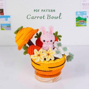 Puede incluir: Un cuenco de zanahoria tejido a ganchillo con un conejito rosa, zanahorias y flores. El cuenco es naranja y amarillo, con una base de madera. El texto "PDF PATTERN Carrot Bowl" es visible.