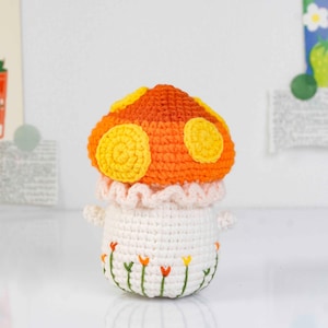 May include: A handmade crochet mushroom toy with an orange and yellow cap, white stem, and small arms. The stem has embroidered flowers. The mushroom has a ruffled pink collar. A whimsical, decorative item.