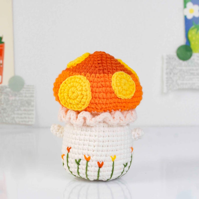 May include: A handmade crochet mushroom toy with an orange and yellow cap, white stem, and small arms. The stem has embroidered flowers. The mushroom has a ruffled pink collar. A whimsical, decorative item.