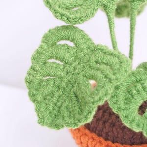 May include: Close-up of a crocheted green plant with large leaves, in a brown and orange pot. The plant is made with a textured yarn and has a realistic appearance.